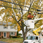 A utility line-clearance crew trimming branches away from overhead power lines using a bucket truck on a suburban residential street.